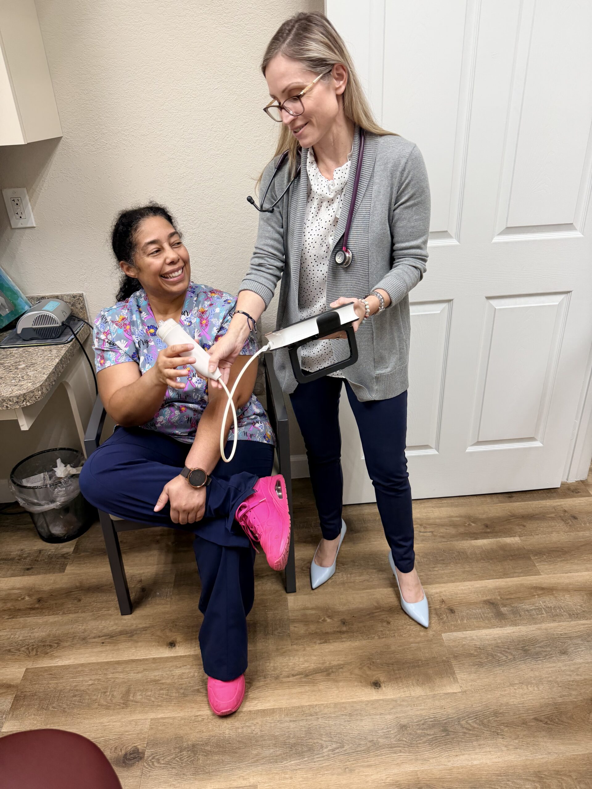 Two women in a clinic, one sitting with a blood pressure cuff, the other standing with a tablet.