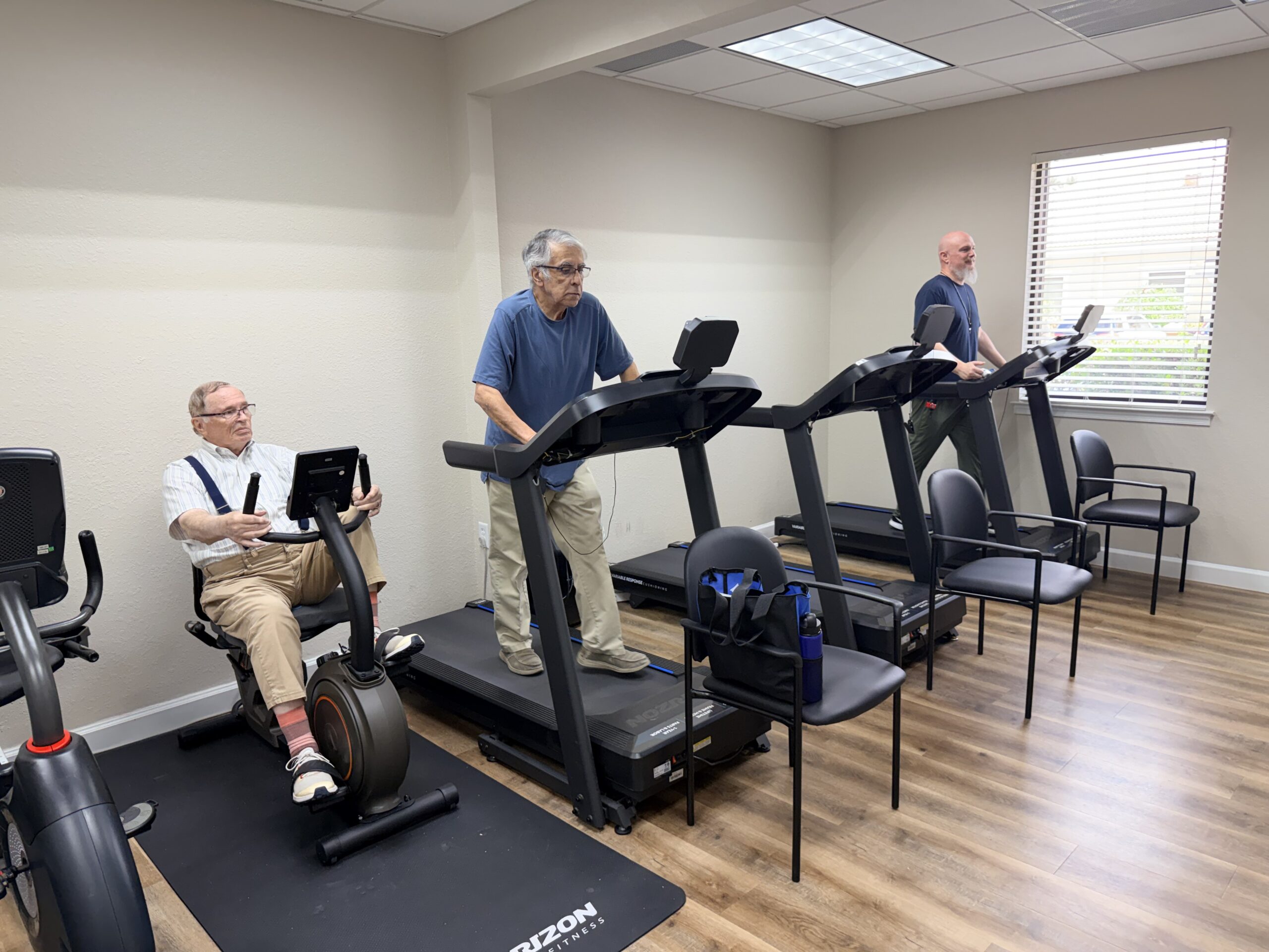 Elderly men exercising on treadmills and a recumbent bike in a gym room.