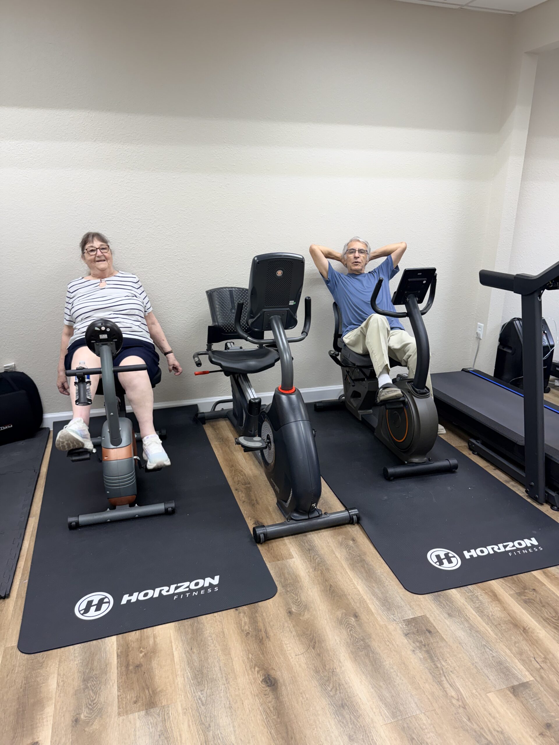 Two elderly people exercising on stationary bikes in a gym.