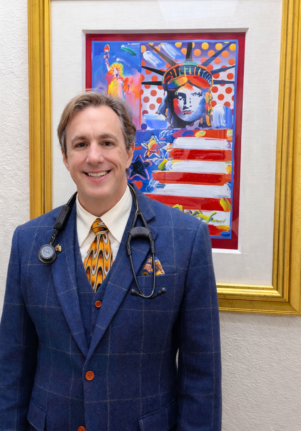 A smiling doctor in a blue suit poses before a colorful Statue of Liberty painting.